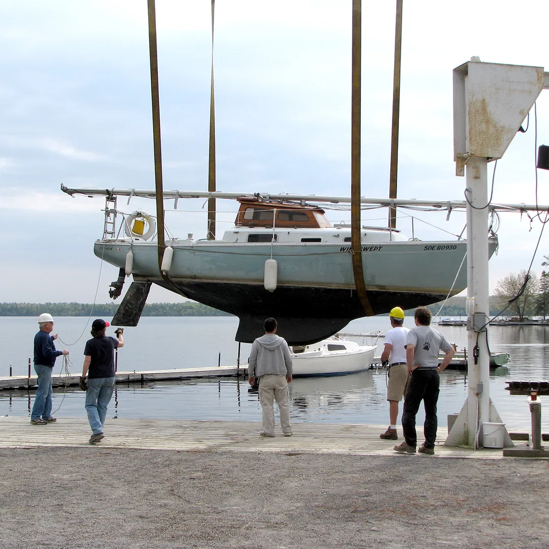 A sailboat is hanging on two straps held by a crane. Five people are standing nearby to guide the boat.